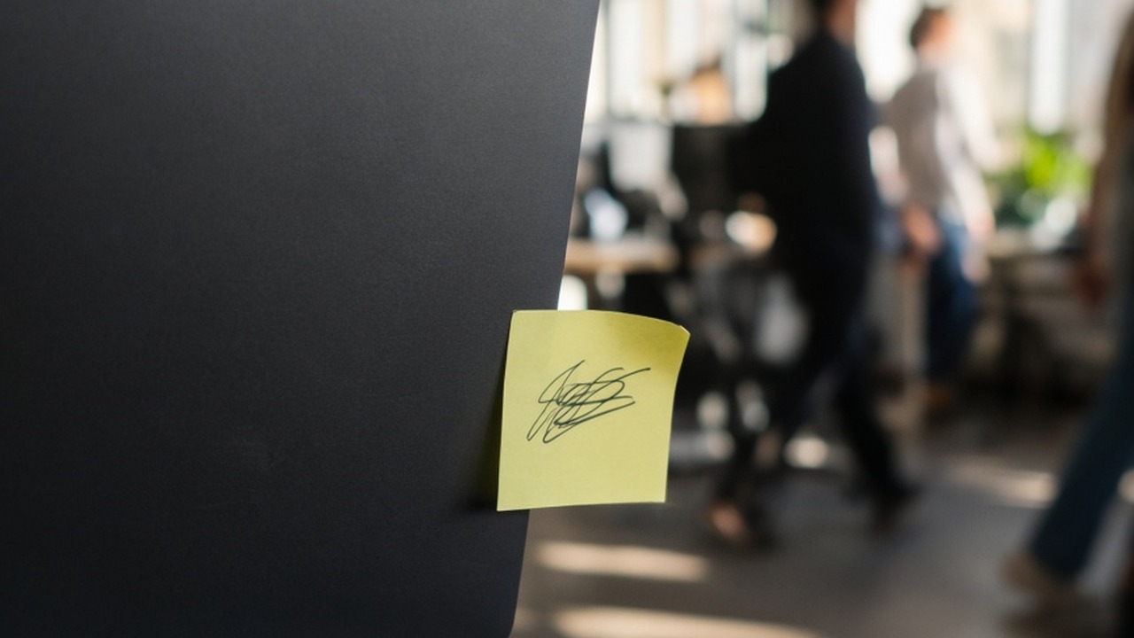 A sticky note with a handwritten password stuck to the edge of a laptop in a busy shared office, people blurred in the background