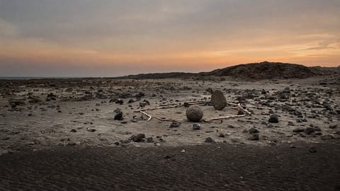 A small deserted island at dusk, with rough stone blocks arranged on the sand into a constellation pattern under a darkening sky.