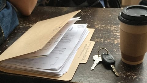 A paper folder marked only with a handwritten title, left open on a cafe counter beside a set of keys and a takeaway coffee, papers half sliding out
