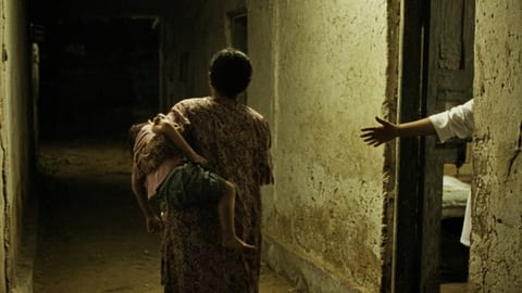 A night clinic corridor in a hot country, a parent carrying a child towards a treatment room, seen from behind.