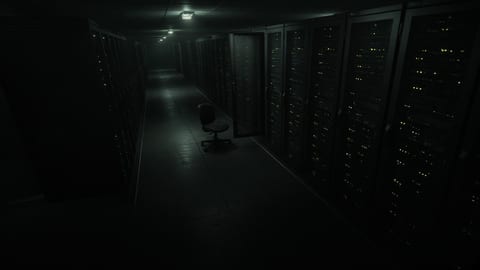 Security-camera style view down a dim, empty data centre aisle at night, one rack door left ajar and an office chair pushed out into the aisle.